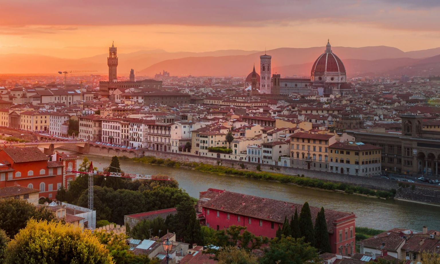 Sunset view of Florence, Italy, showcasing the Arno River, historic buildings, the iconic Florence Cathedral, and the Palazzo Vecchio tower.