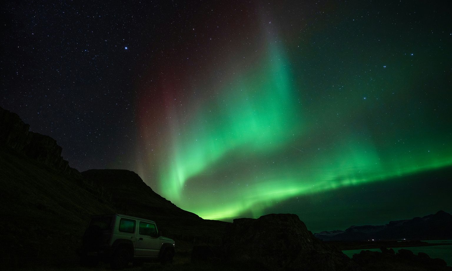 Northern lights with green and red hues illuminate a starry night sky above a rugged landscape with a parked SUV in the foreground.