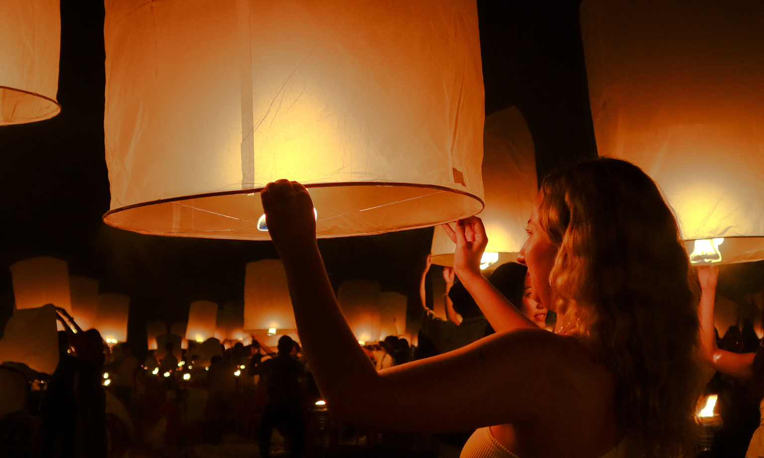A woman releases a glowing paper lantern into the night sky, surrounded by others doing the same, creating a warm, illuminated scene.