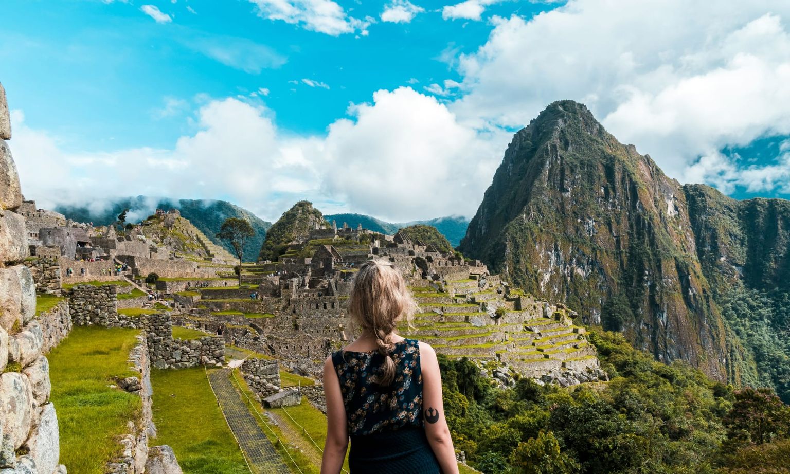 A woman with blonde hair stands overlooking the ancient ruins of Machu Picchu, surrounded by green mountains under a partly cloudy sky.