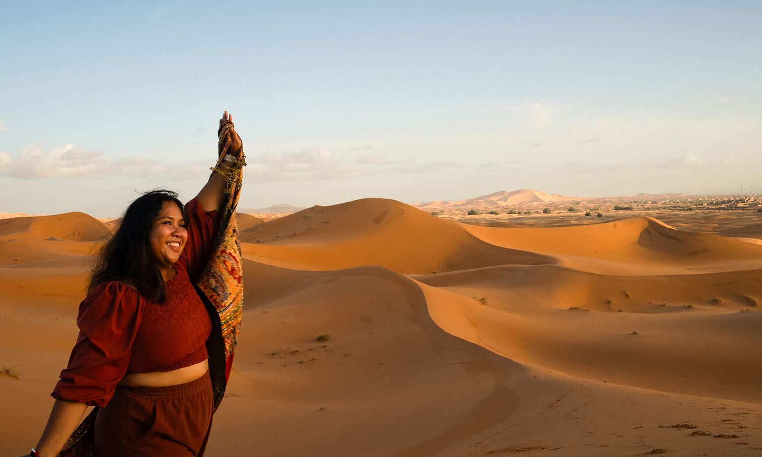 A woman in a red outfit joyfully raises her hand against a backdrop of vast, sunlit sand dunes under a clear blue sky.