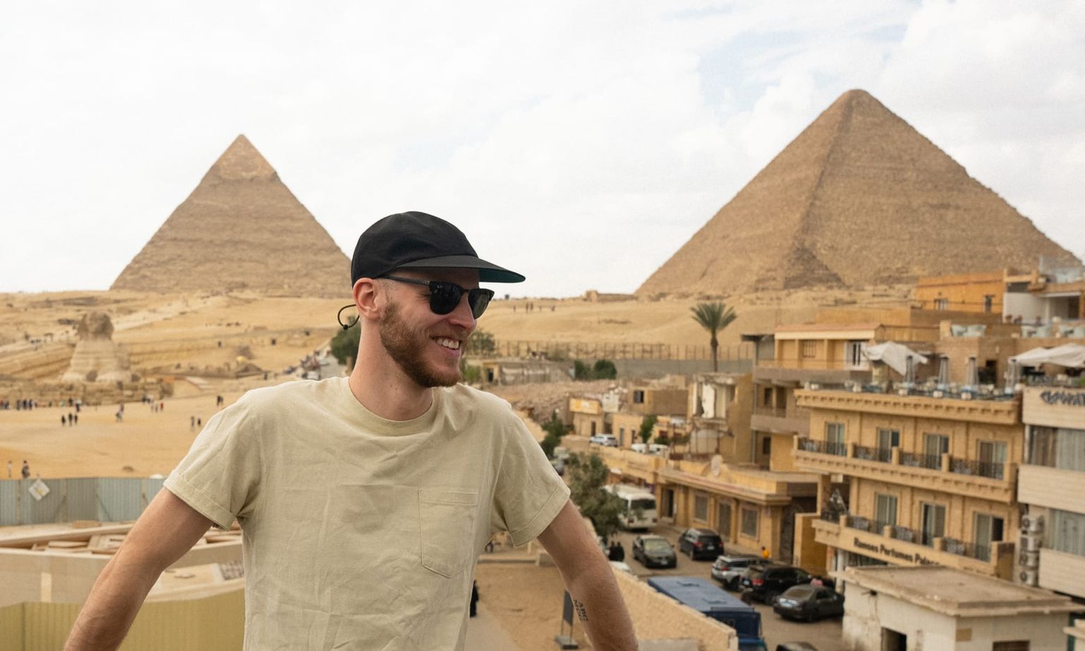 Man in sunglasses and a cap smiles with the Pyramids of Giza in the background, under a partly cloudy sky.