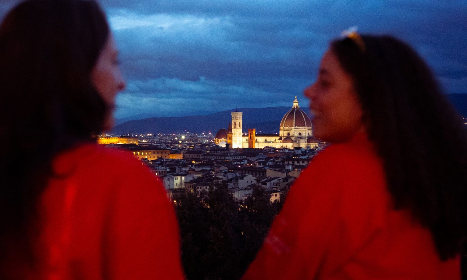 Two people in red jackets look at a cityscape featuring a lit-up dome and tower at dusk, with dramatic clouds in the sky.