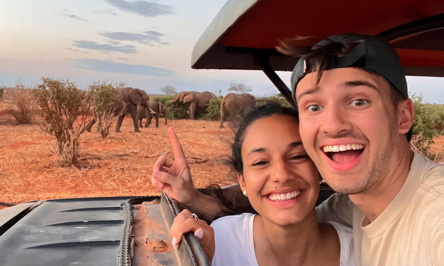 A smiling couple takes a selfie on a safari with elephants in the background, standing next to their vehicle on a sunny day.