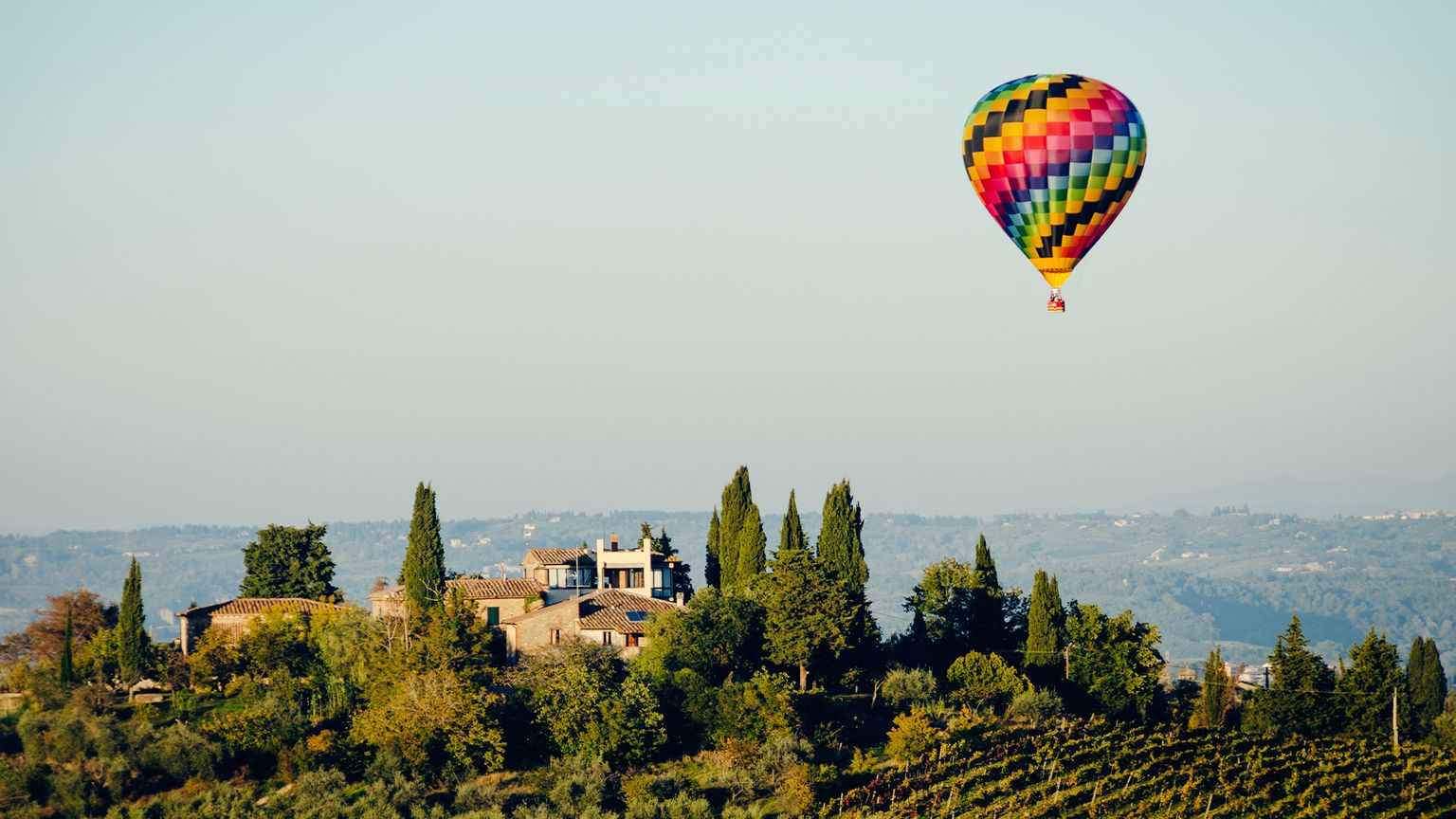 A coloful hot air balloon floating over a home on a tree-covered hillside and vineyard in Tuscany, Italy
