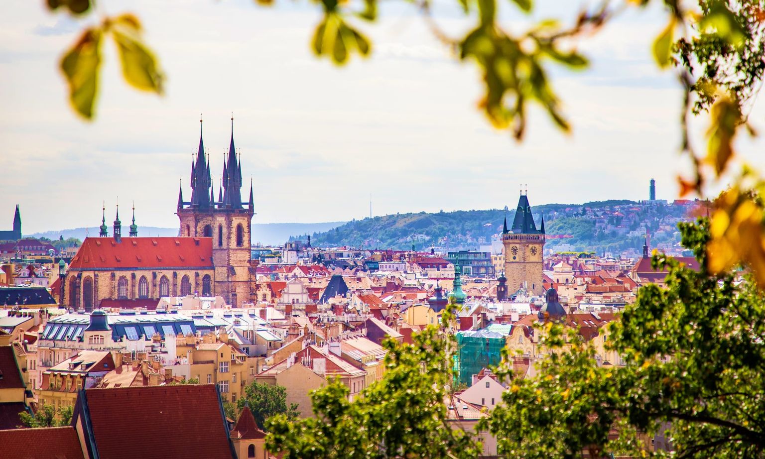 Scenic view of Prague's skyline with historic buildings, prominent church spires, and greenery in the foreground.