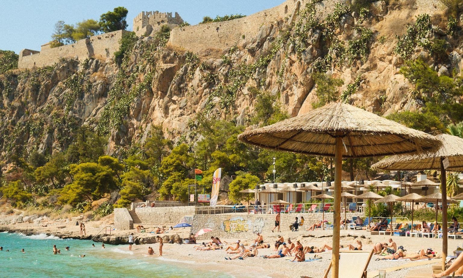A sunny beach with people sunbathing and swimming, set against rocky cliffs with a historic stone fort on top.