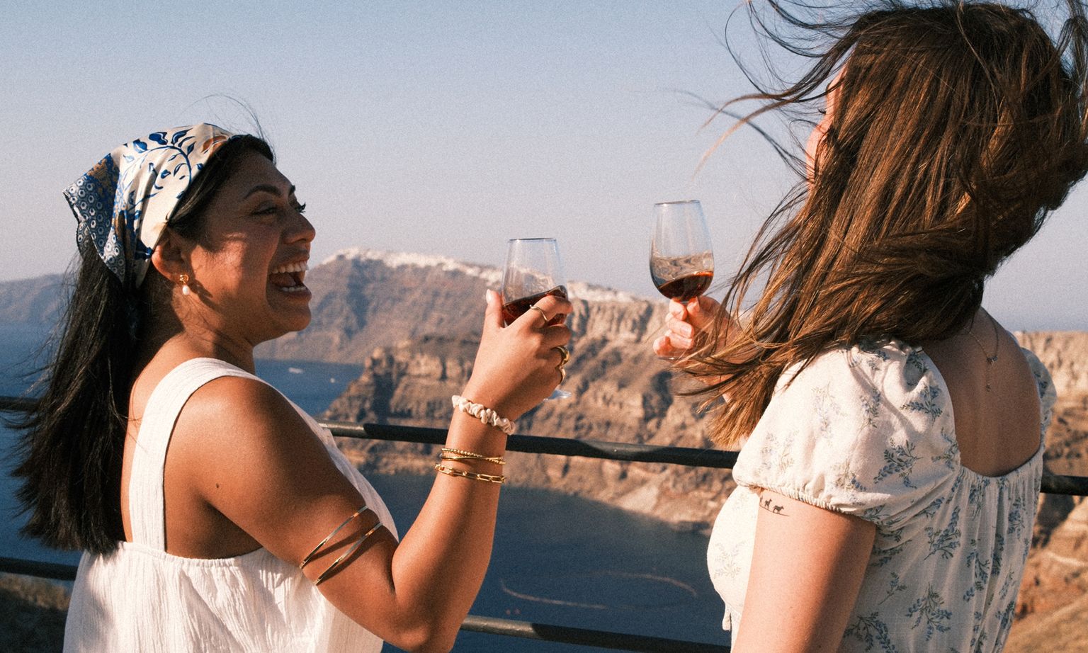 Two women joyfully toast with wine glasses on a scenic balcony overlooking a rocky coastline, with windblown hair and vibrant outfits.