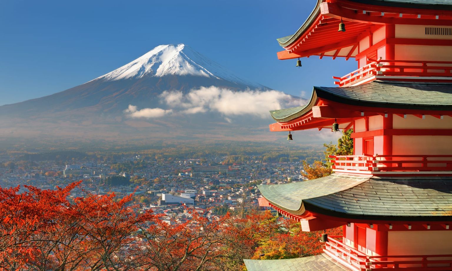 Red pagoda with green roof in foreground, vibrant autumn trees, and Mount Fuji capped with snow under a clear blue sky in the background.