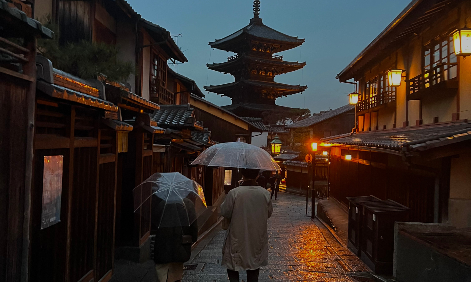 Two people with umbrellas walk down a lantern-lit street in Kyoto, Japan, with a traditional pagoda in the background at dusk.