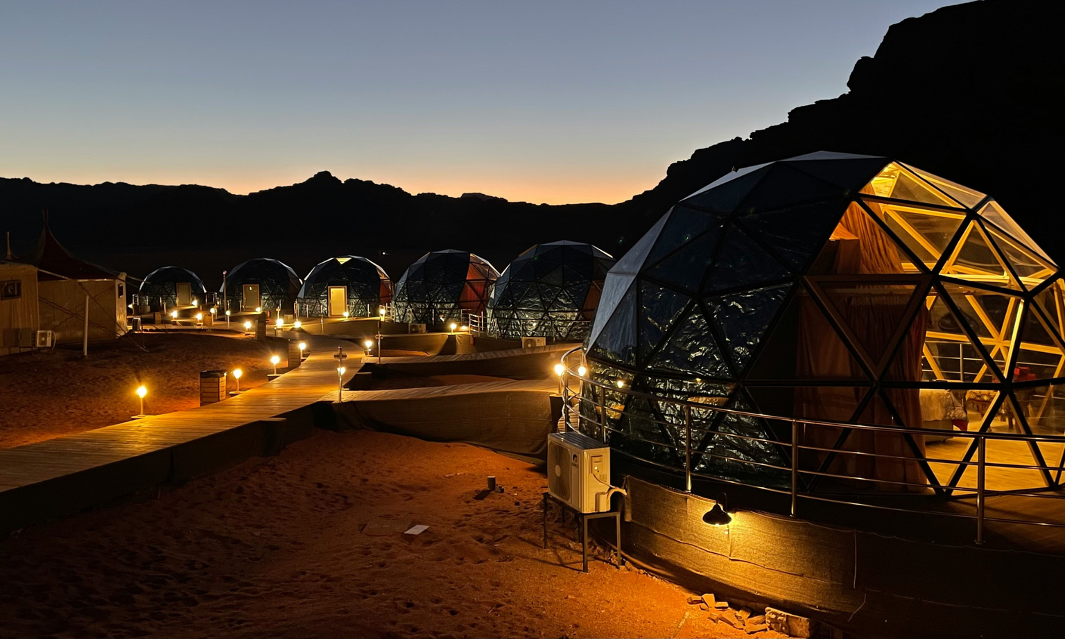 A row of illuminated geodesic dome tents on a desert landscape at dusk, with a wooden walkway and a mountainous silhouette in the background.