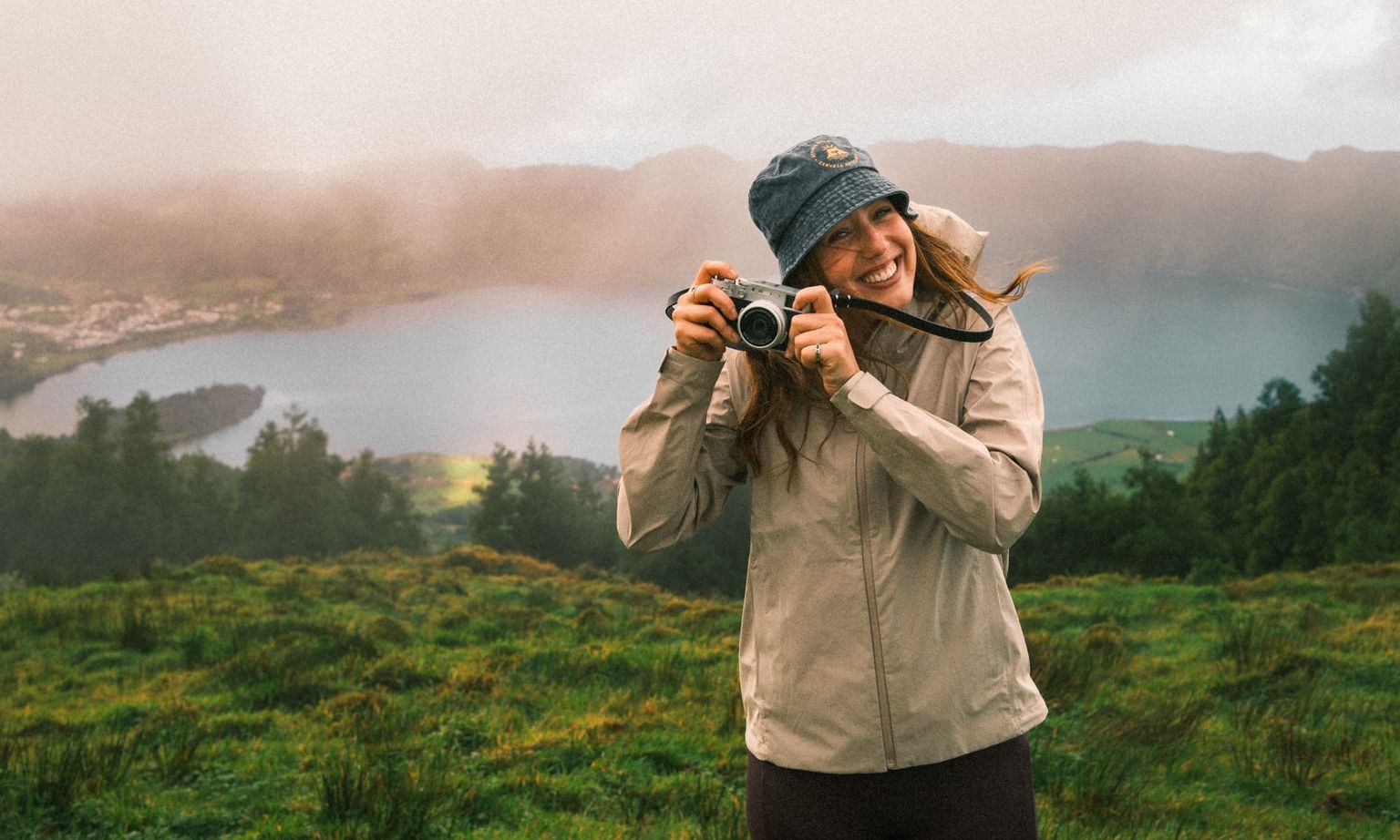 Woman in a light jacket and bucket hat smiling, holds a camera with a scenic lake and cloudy sky in the background.