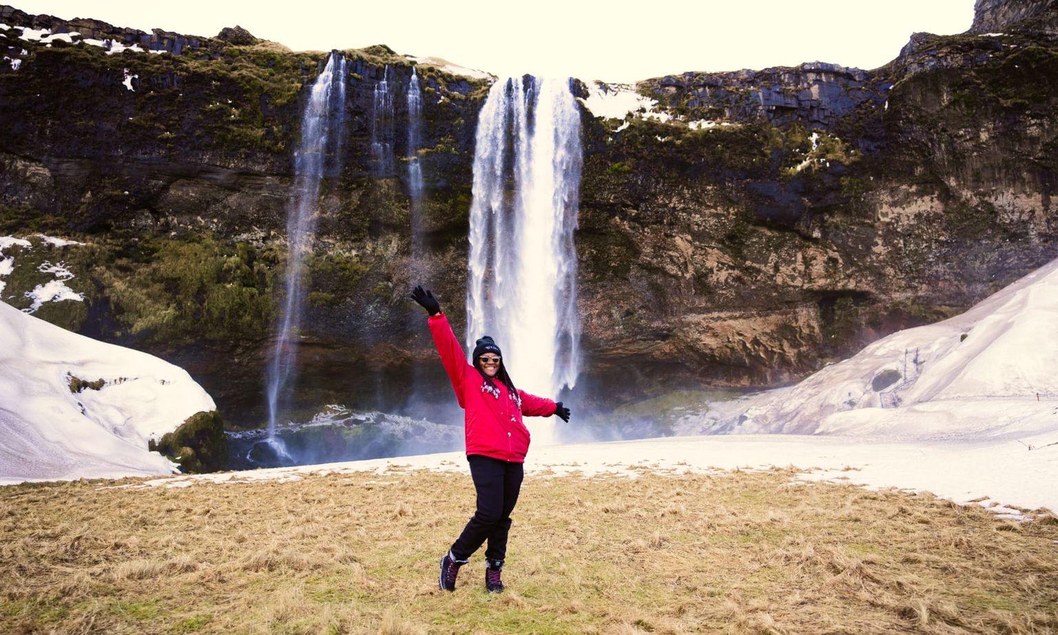 Person in a red jacket joyfully poses in front of a tall waterfall cascading down a rocky cliff surrounded by snowy patches.