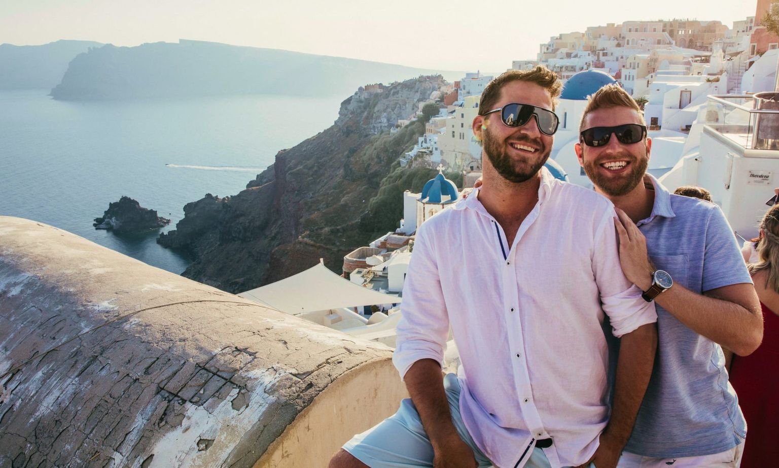 Two smiling men wearing sunglasses pose together on a sunny cliffside in Santorini, Greece, with white buildings and the sea in the background.