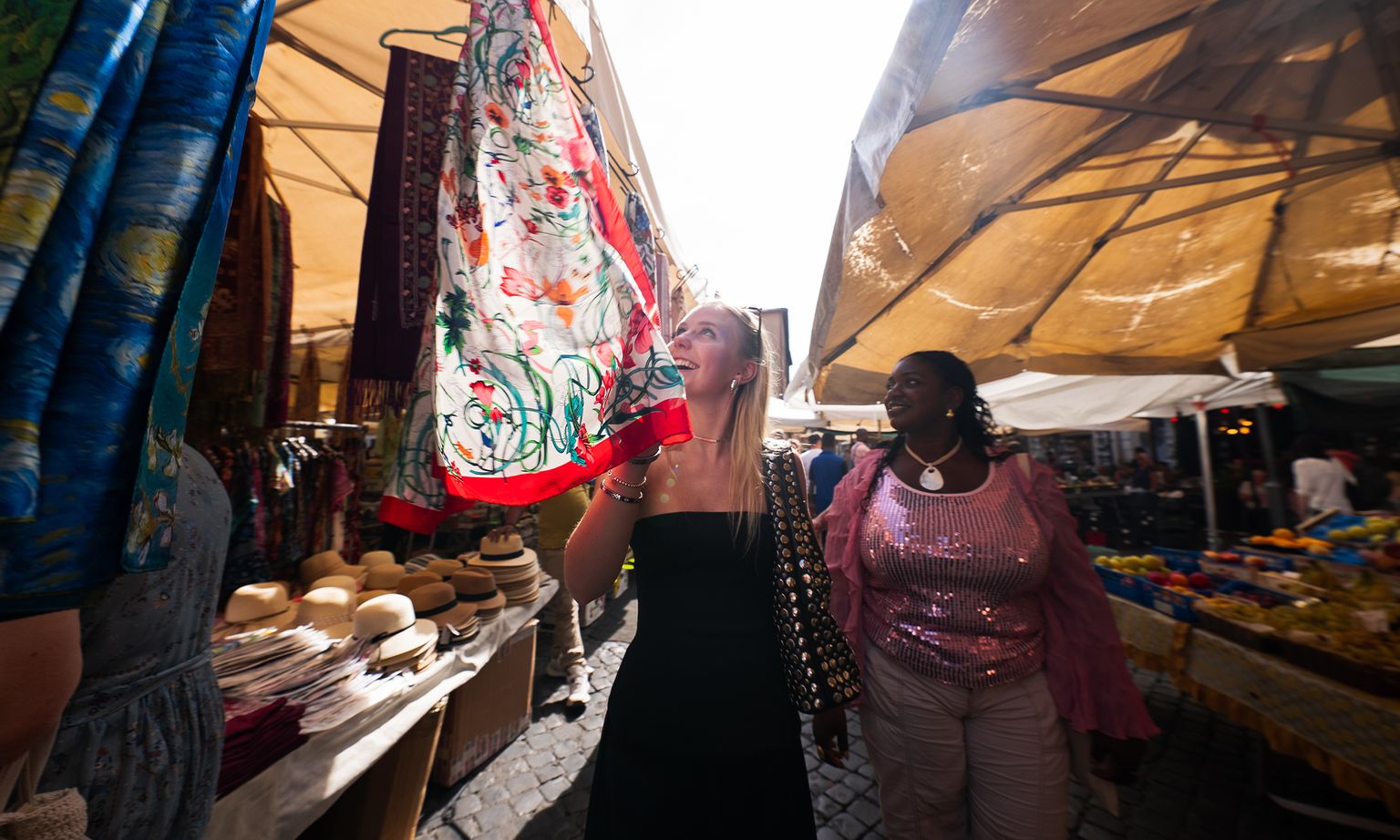 Two women shopping at an outdoor market, admiring colorful fabric hanging under a canopy, with woven hats displayed on a table nearby.