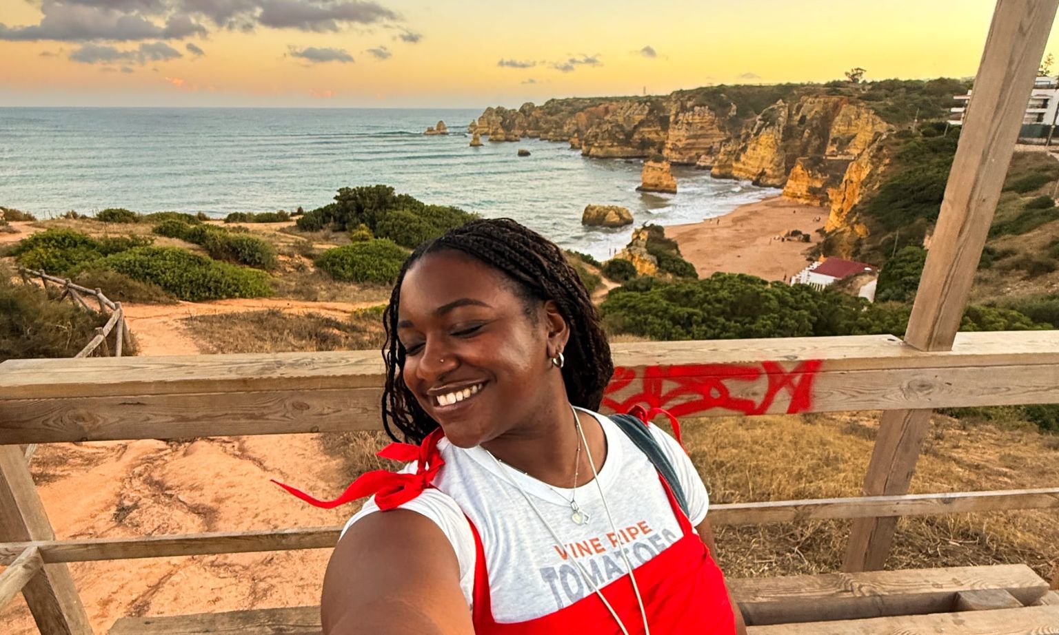 Woman smiling for a selfie on a wooden deck overlooking a scenic coastal view with cliffs, beach, and ocean under a cloudy sky.
