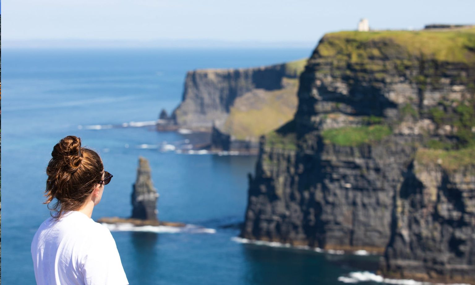 Person with a bun gazes at the Cliffs of Moher, overlooking the ocean and rocky cliffs under a clear blue sky.