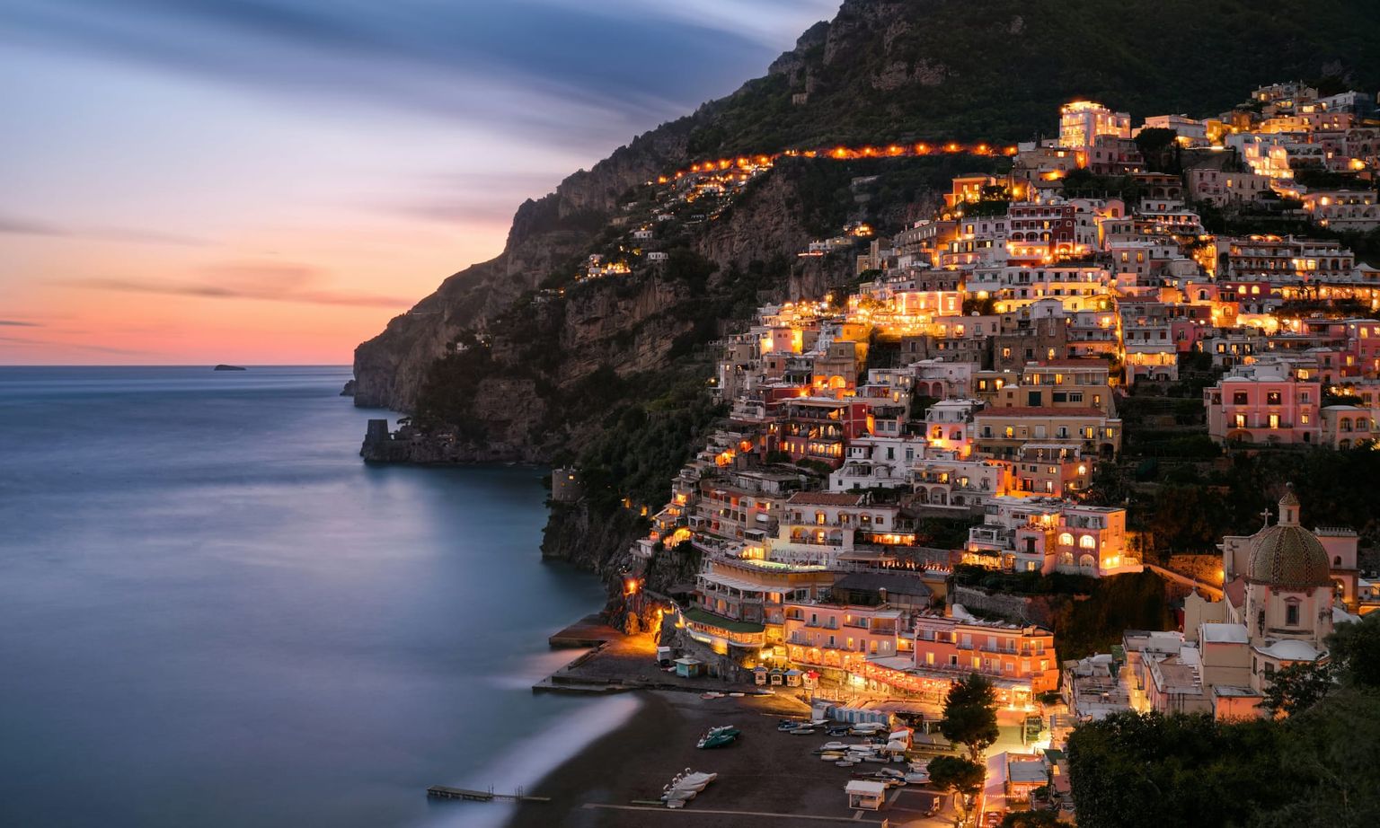 Scenic view of a coastal village at dusk, with illuminated buildings cascading down a hillside towards a calm sea under a colorful sky.