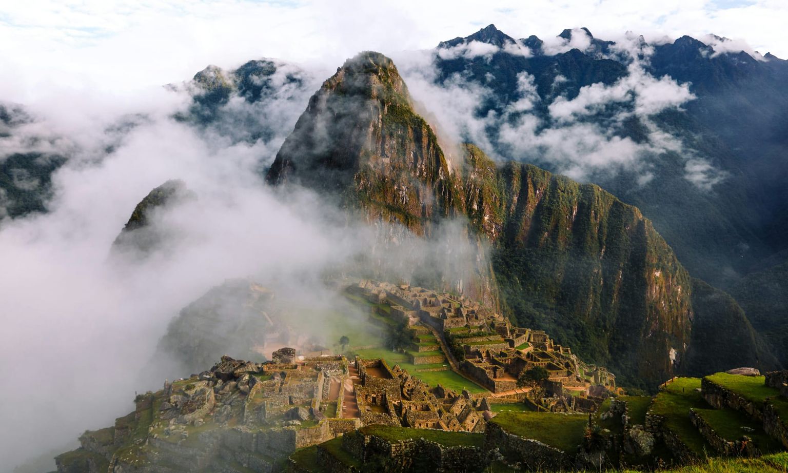 Machu Picchu shrouded in mist, surrounded by steep mountains and lush vegetation under a partly cloudy sky.