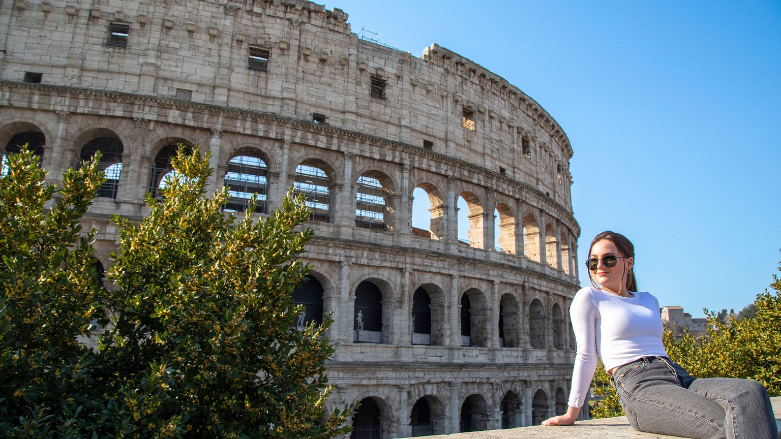 A person wearing sunglasses sits on a ledge with the Colosseum in the background on a sunny day.
