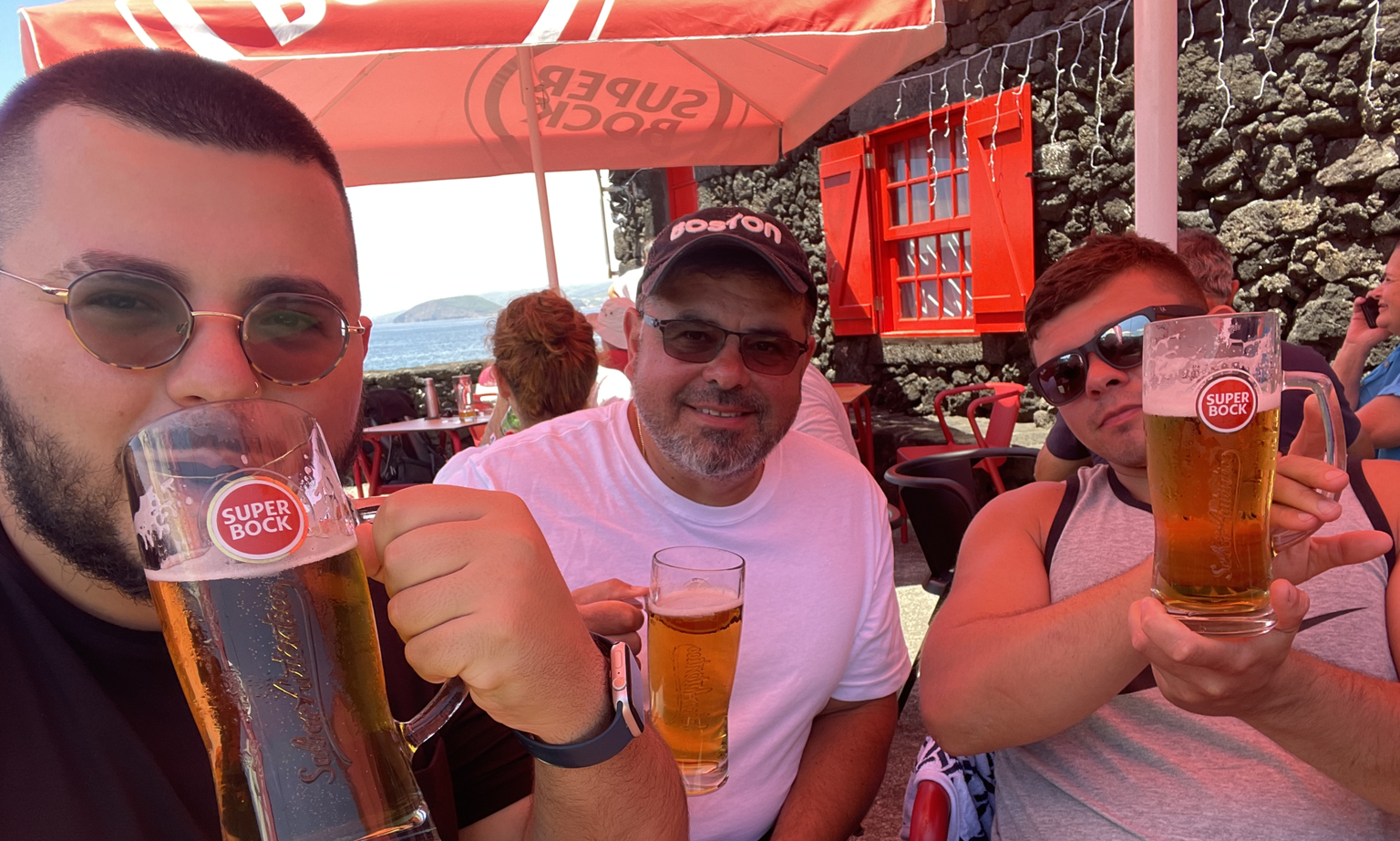 Three men smiling and holding steins of beer sitting at an outdoor. cafe