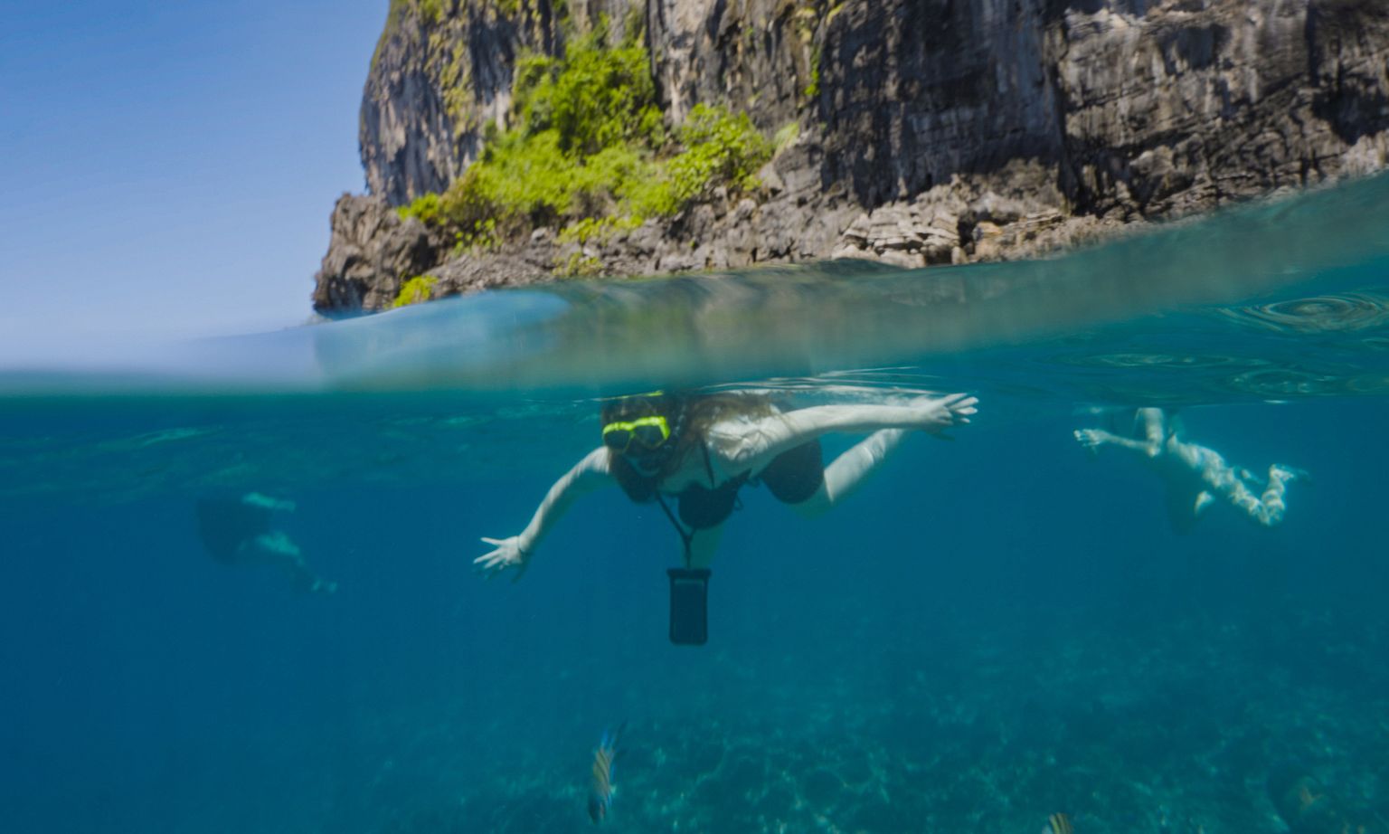 A person snorkeling in clear blue water near a rocky cliff with greenery; two others swim nearby.