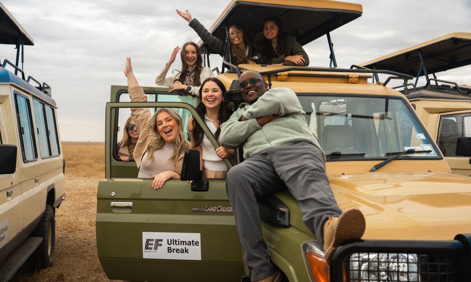A group of smiling people pose enthusiastically on and around a safari vehicle in an open, grassy landscape.