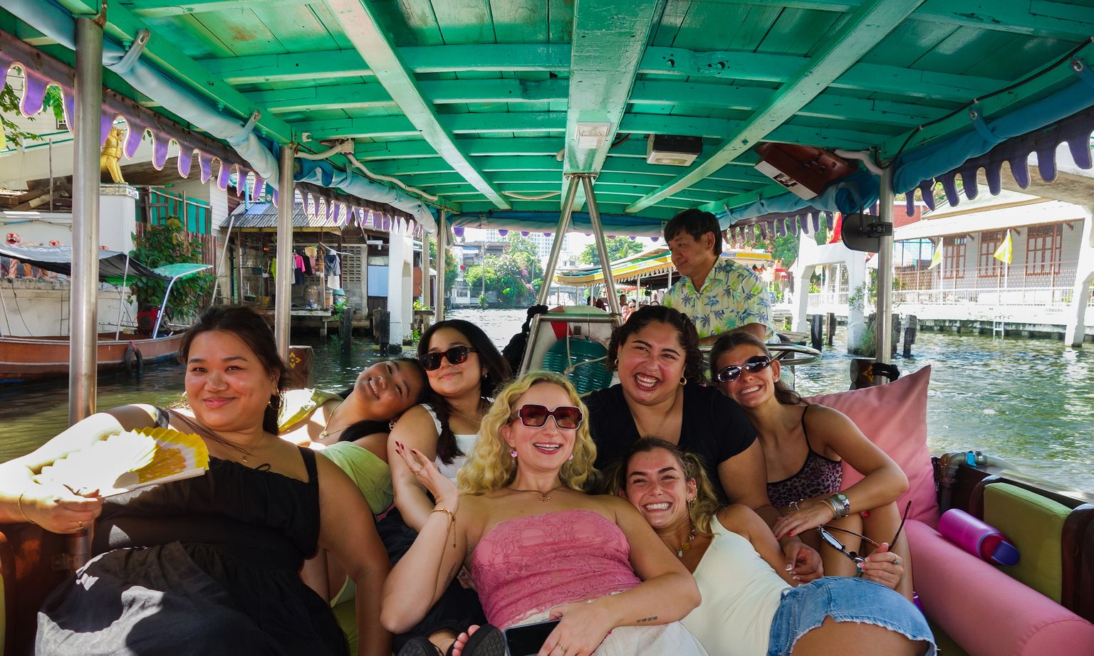 A group of smiling people relax on a colorful boat, cruising along a canal with vibrant buildings in the background.