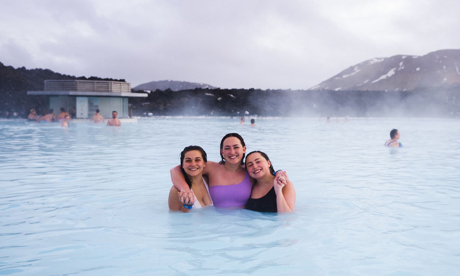Three people smiling and embracing in a steaming outdoor hot spring, with misty mountains in the background.