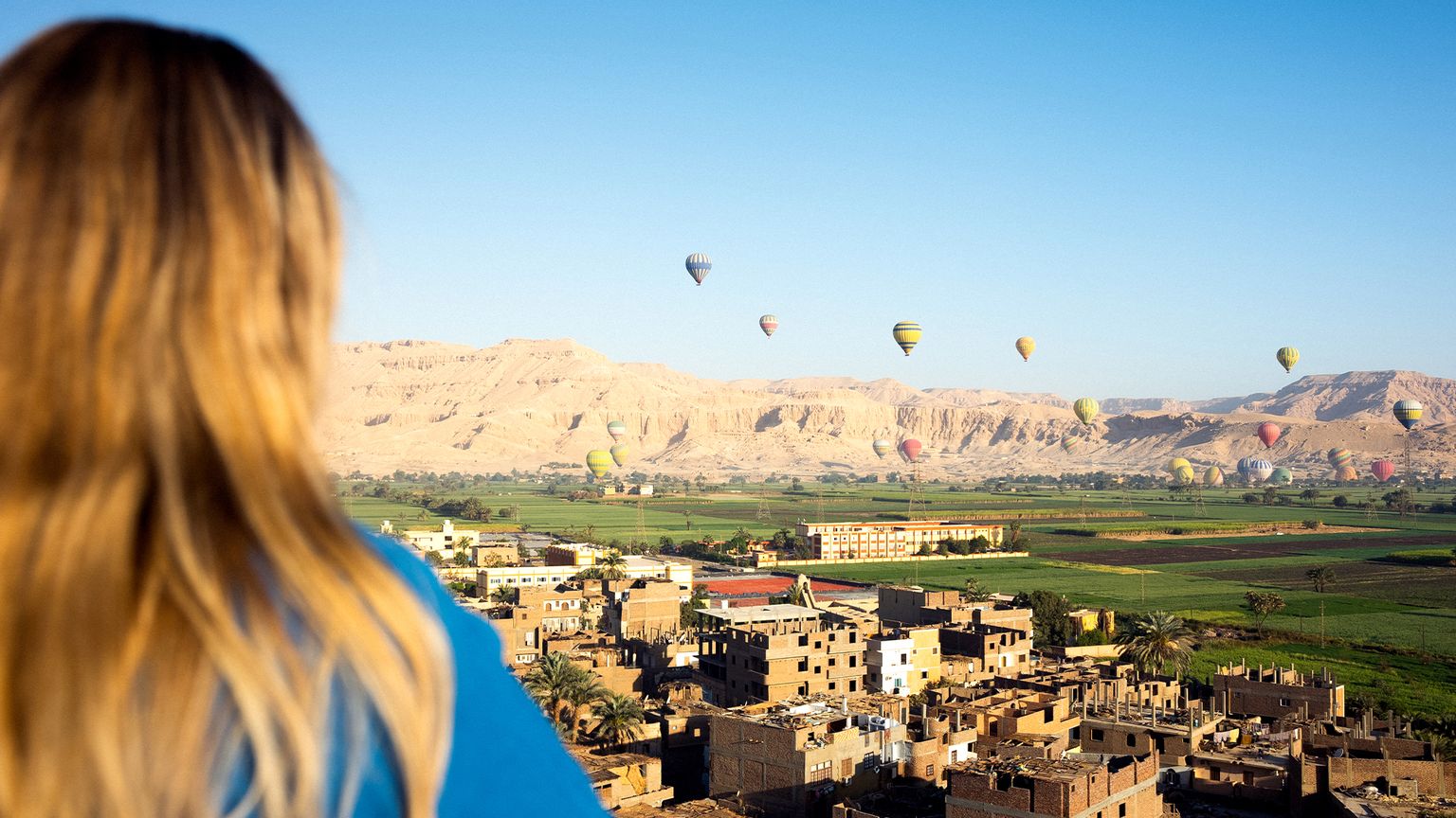 A person looking out toward many hot air balloons floating over the buildings, green fields, and mountains of Luxor, Egypt