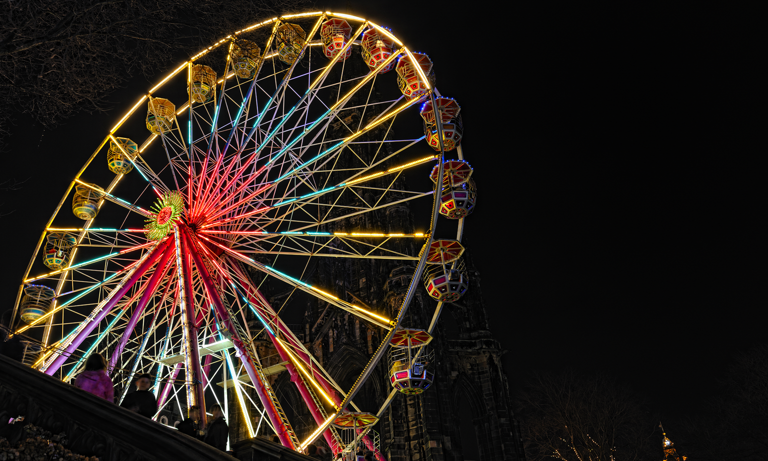 A brightly lit Ferris wheel at night, with colorful cabins and illuminated spokes against a dark sky. Silhouetted trees frame the left.
