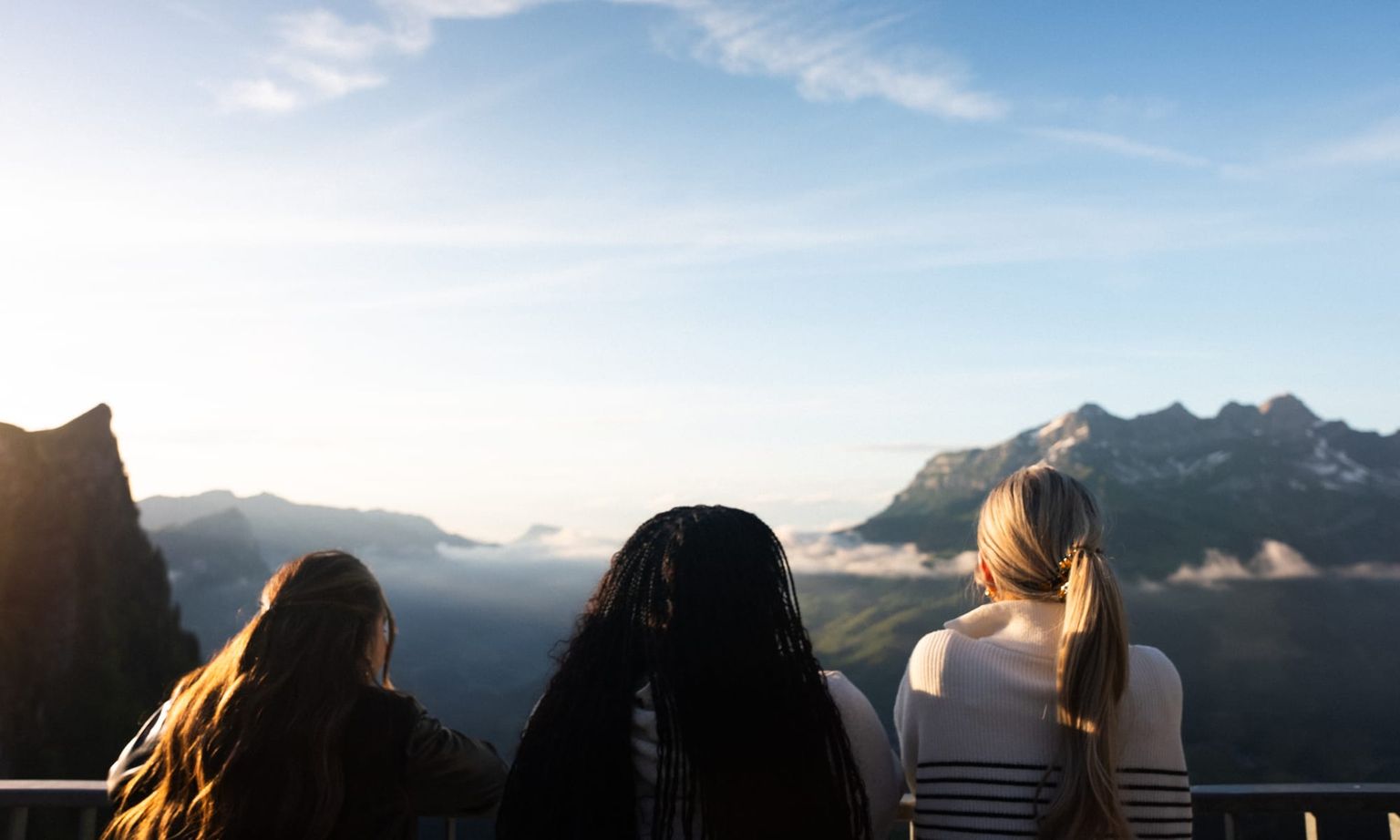Three people with long hair stand on a balcony, overlooking a vast mountain landscape under a clear, blue sky.