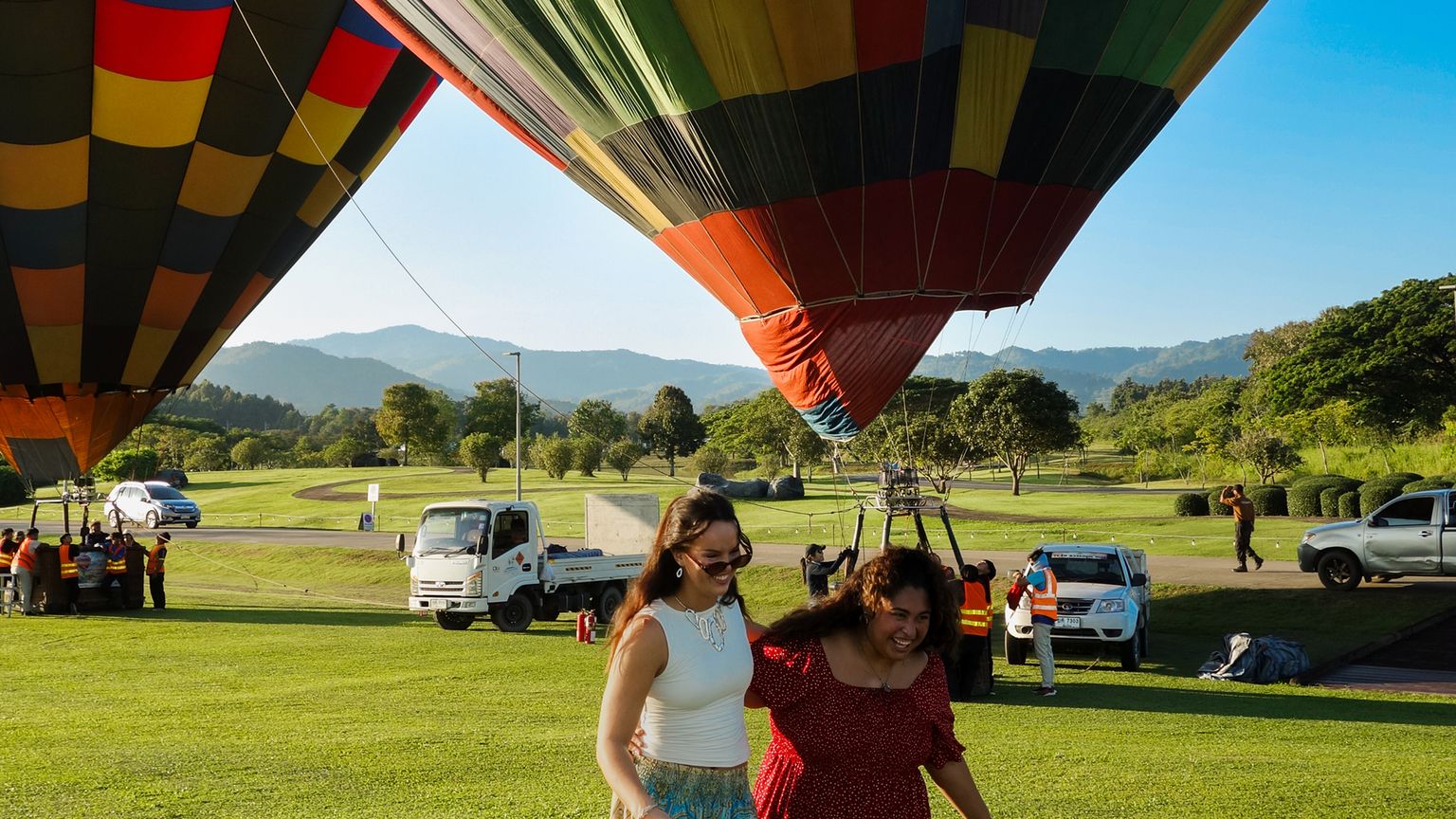 Two women smiling and walking in front of colorful hot air balloons being prepared for flight in a scenic, grassy area with mountains in the background.