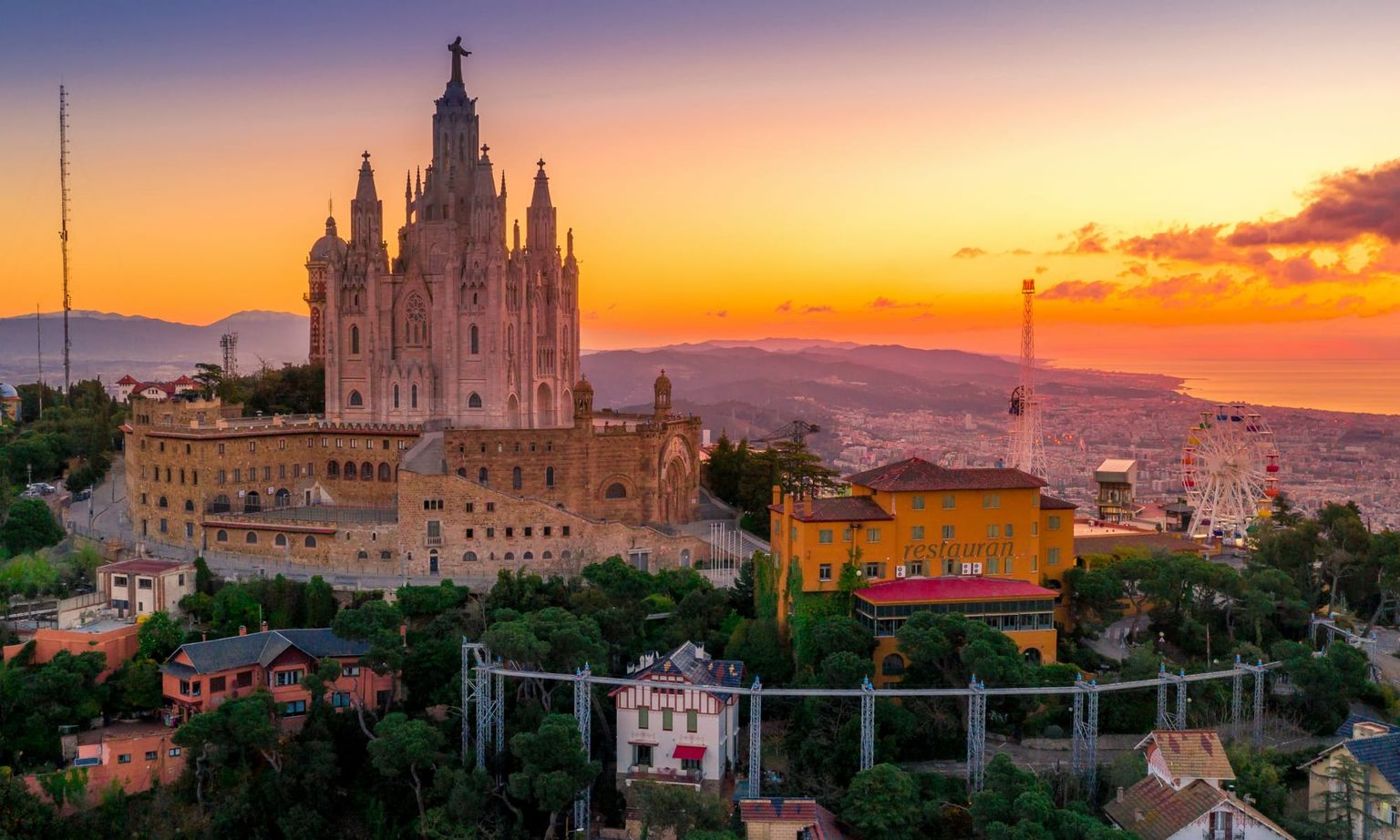 Sunset view of Tibidabo Amusement Park and the Temple of the Sacred Heart of Jesus in Barcelona, with a Ferris wheel and the city skyline.