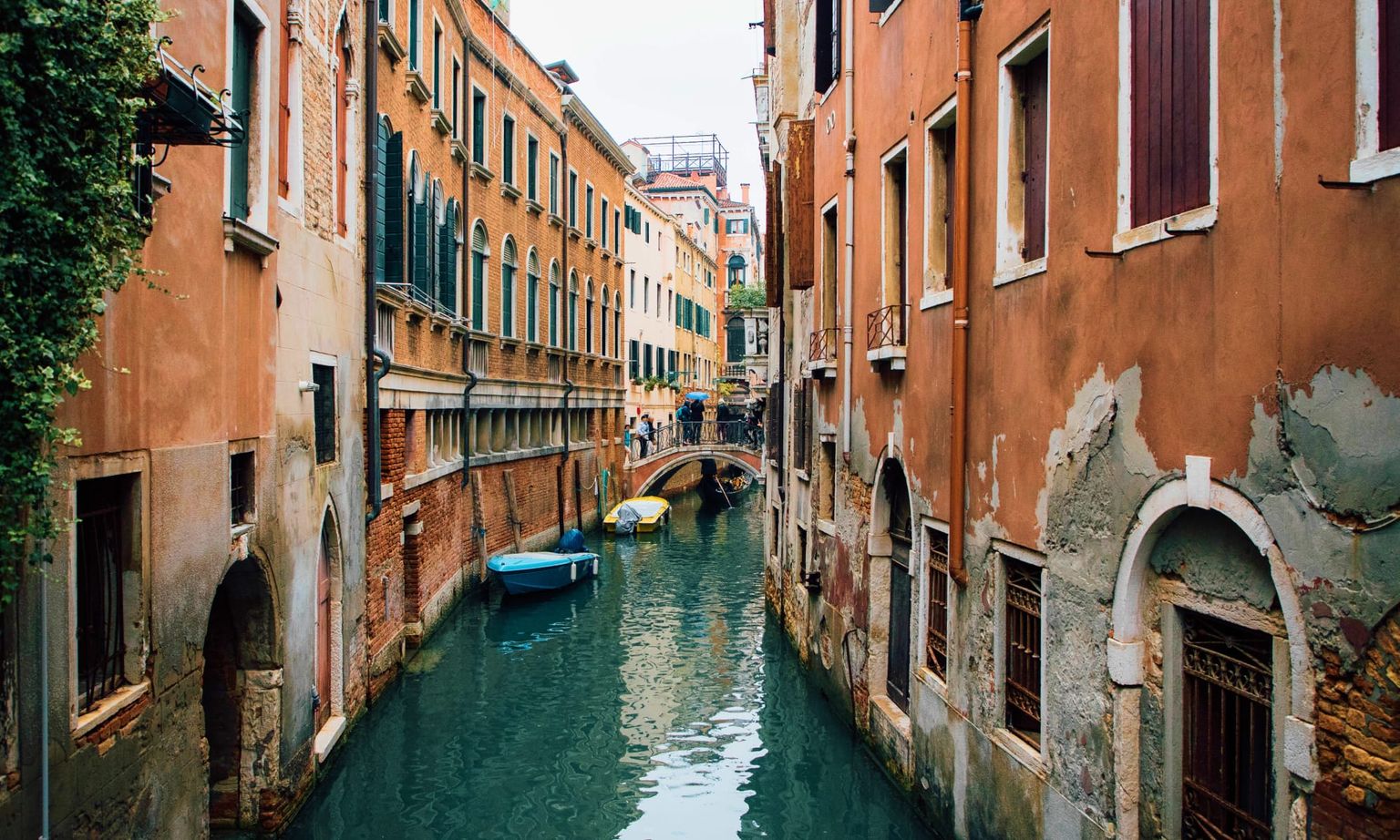 Narrow Venetian canal with calm water, colorful boats, and historic buildings on both sides. A small footbridge connects the buildings.