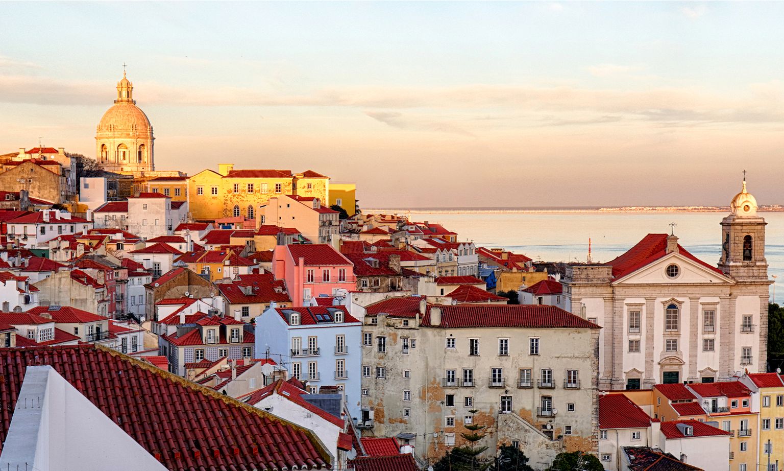 Colorful, red-roofed buildings and a domed church on a hill overlooking the Tagus River in Lisbon, Portugal