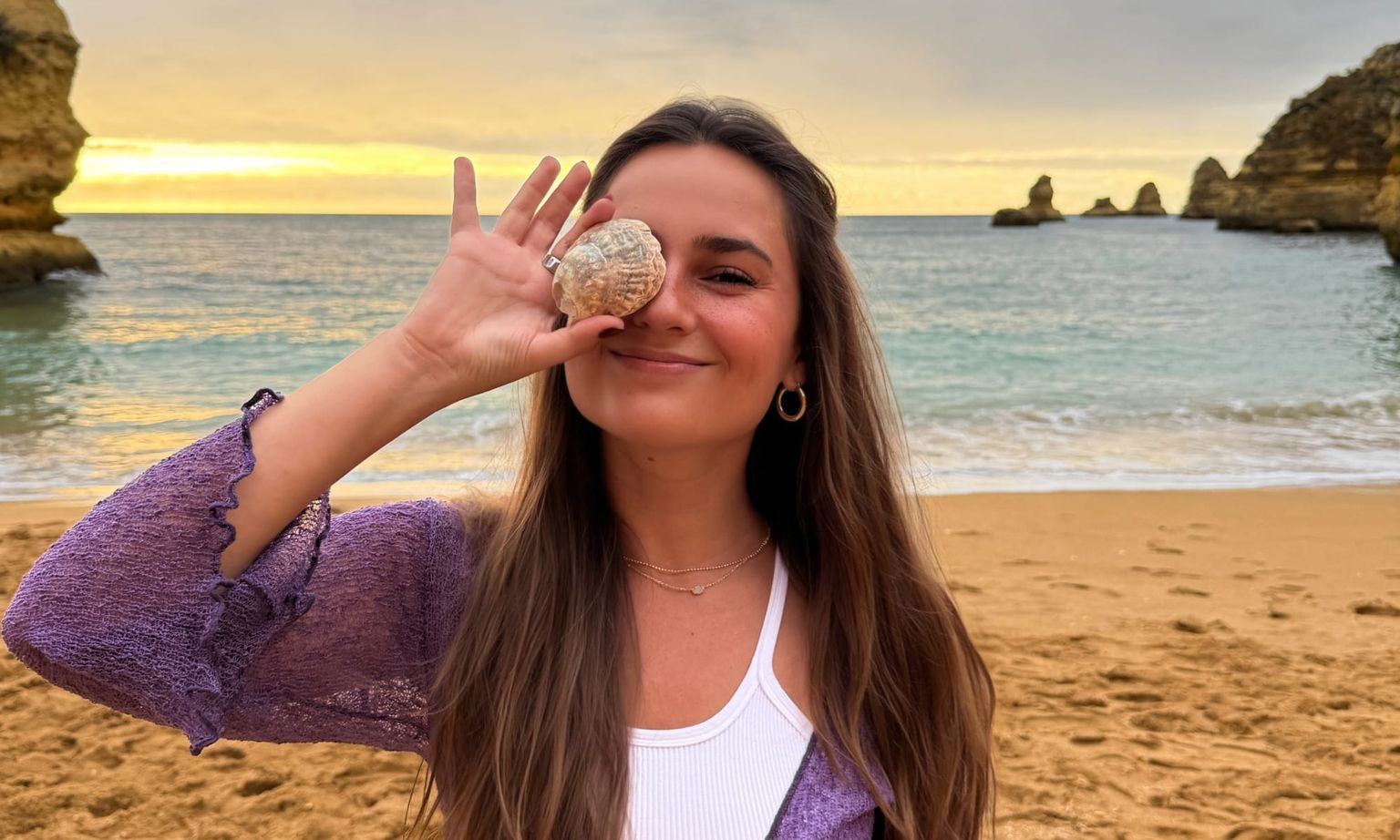 Woman smiling on a beach at sunset, holding a seashell over one eye. Rocky formations and calm sea in the background.
