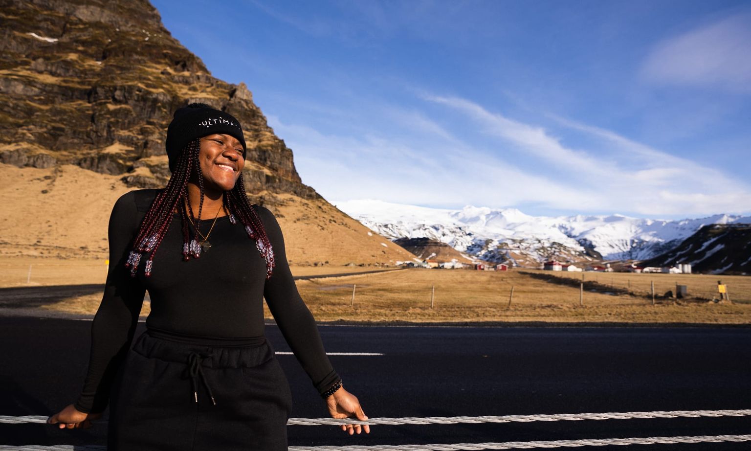 Smiling person in black outfit stands by a road, with a backdrop of mountains under a clear blue sky.