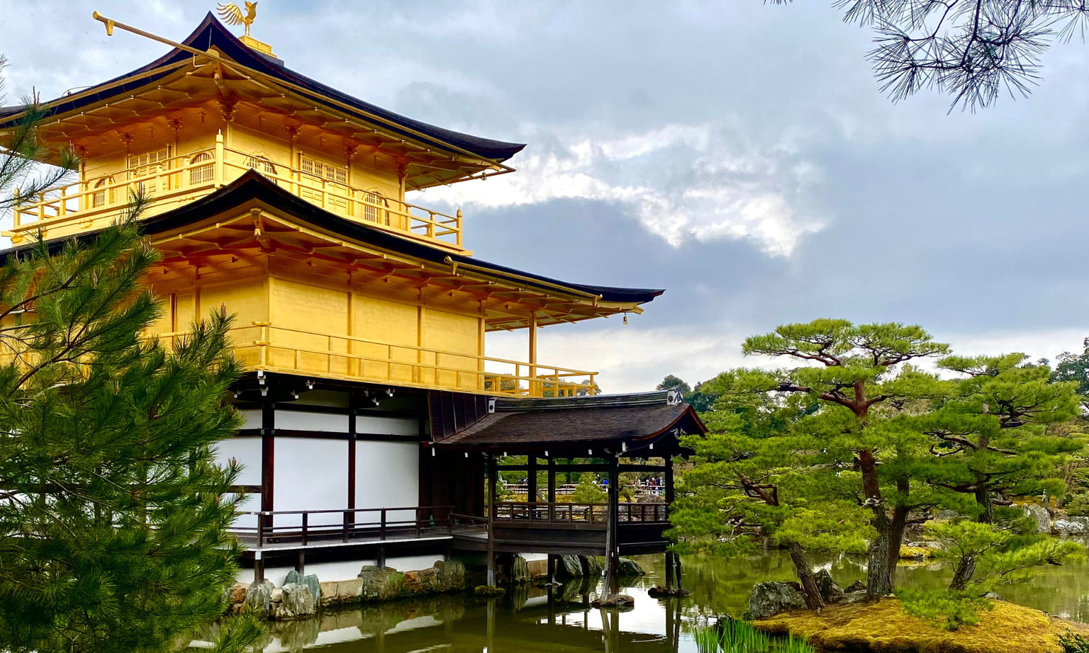 Golden pavilion surrounded by trees and a reflective pond under a cloudy sky.