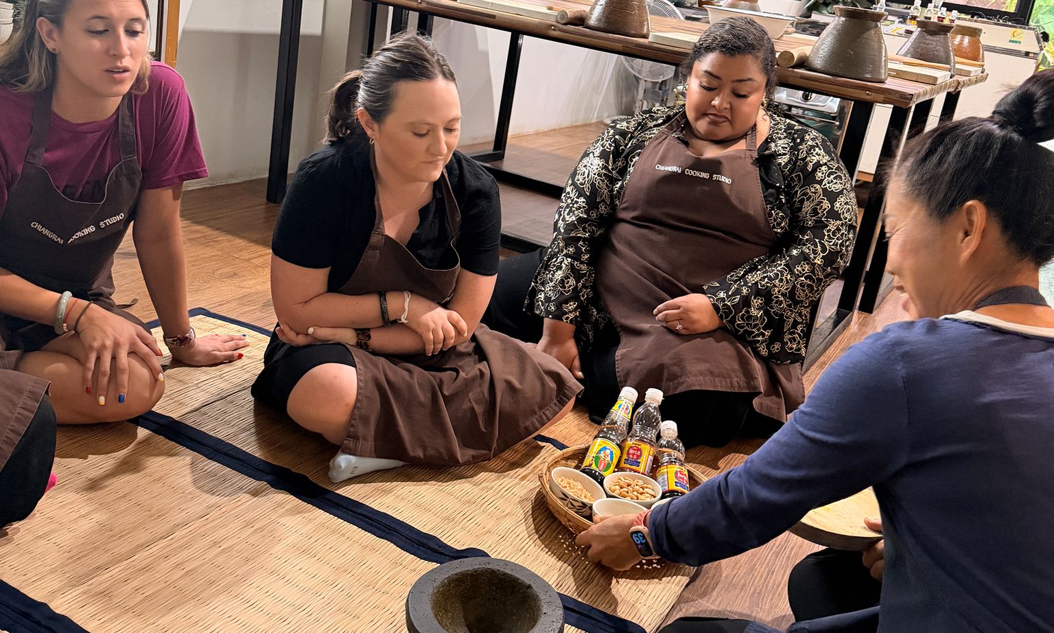 People sitting on mats around a wooden tray with condiments and snacks, engaged in a food preparation activity, wearing aprons.