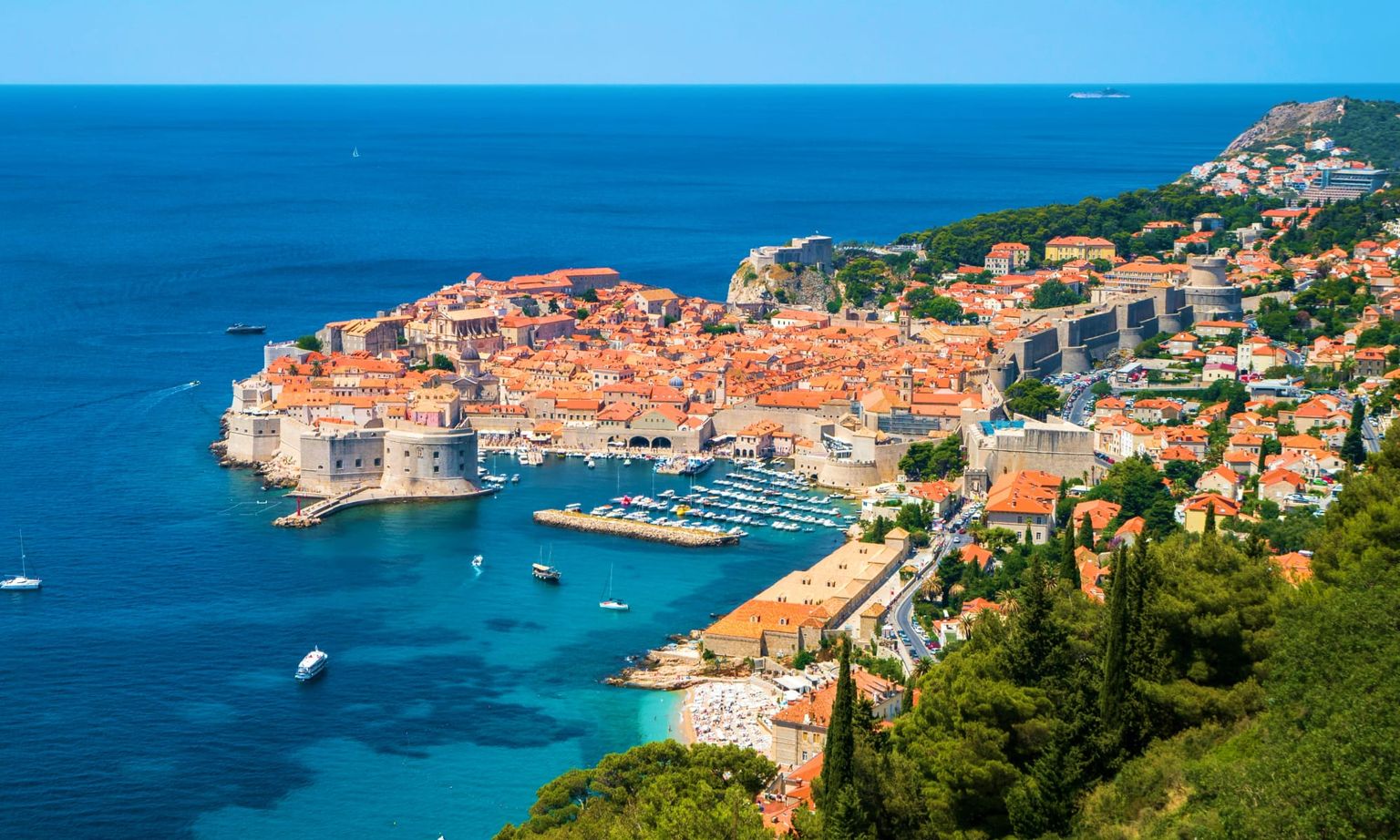 Aerial view of Dubrovnik, Croatia, featuring historic buildings with orange roofs, a harbor, and the surrounding Adriatic Sea.