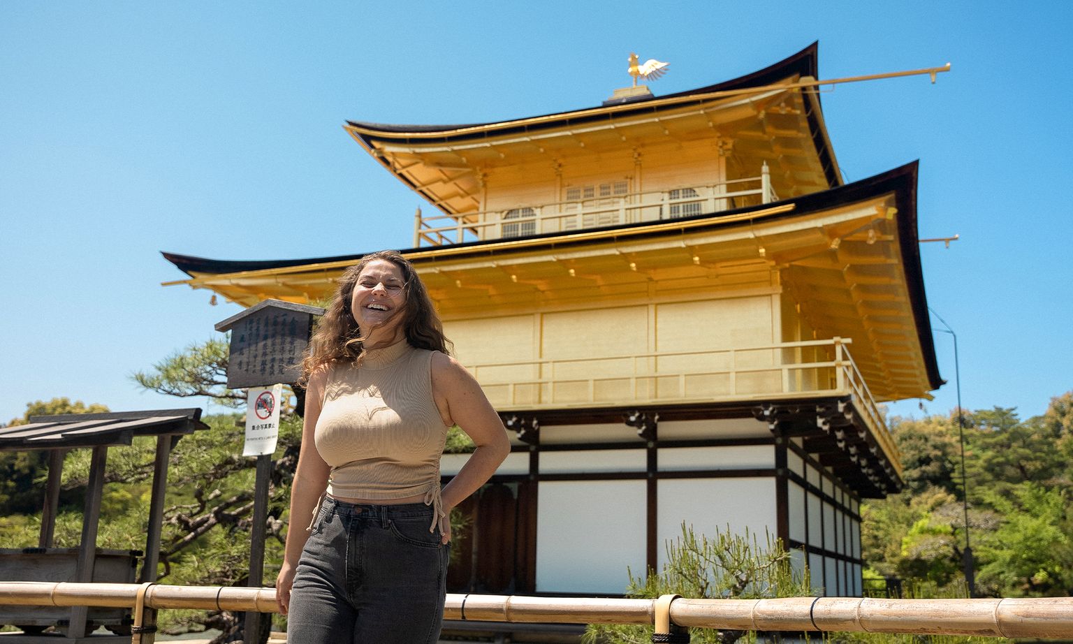 A woman smiles near a traditional Japanese building with golden accents, set against a clear blue sky and surrounded by greenery.