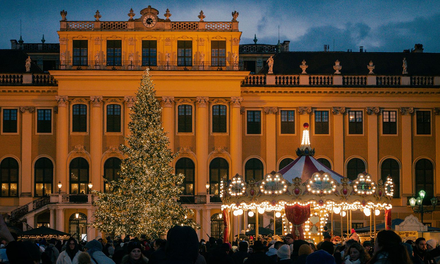 A festive scene with a decorated Christmas tree and carousel in front of a lit, ornate building at dusk. Crowds gather in the foreground.
