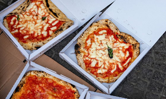 Three margherita pizzas in open boxes on a textured surface, each topped with melted mozzarella and tomato sauce.