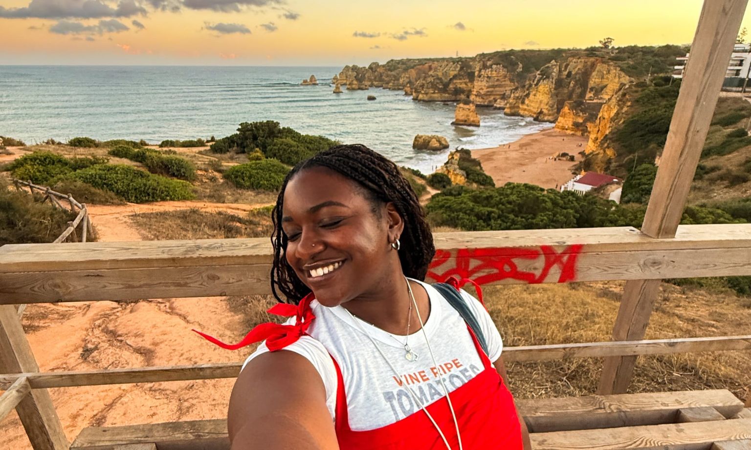 Woman smiling for a selfie with cliffs, ocean, and a sandy beach in the background during sunset.