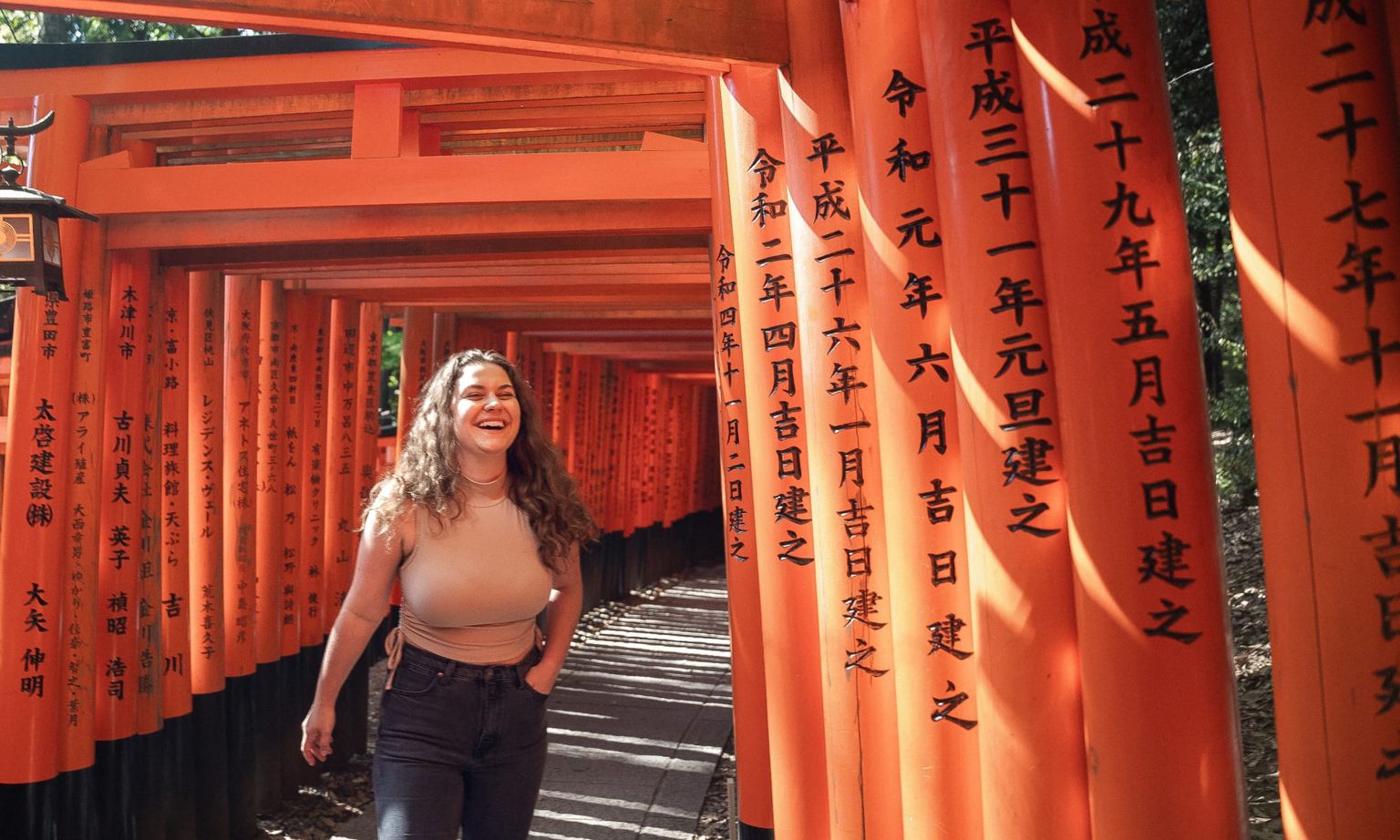 A person joyfully walks through a vibrant orange torii gate tunnel, with sunlight casting shadows. Japanese characters adorn the gates.