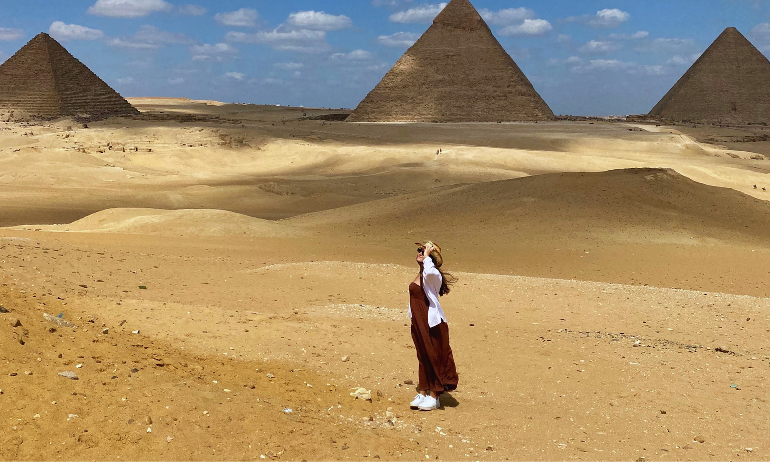 Person in brown dress and hat standing in desert with pyramids in the background under a blue sky with clouds.