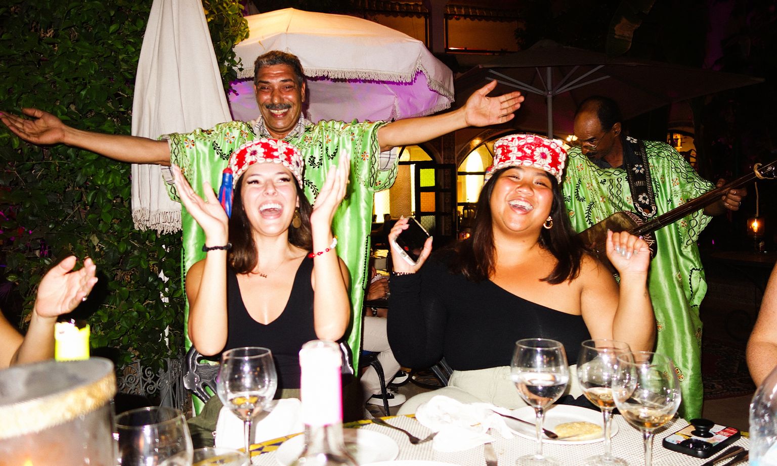 Two women laughing and wearing decorative hats at a lively outdoor dinner, with musicians in green costumes performing in the background.