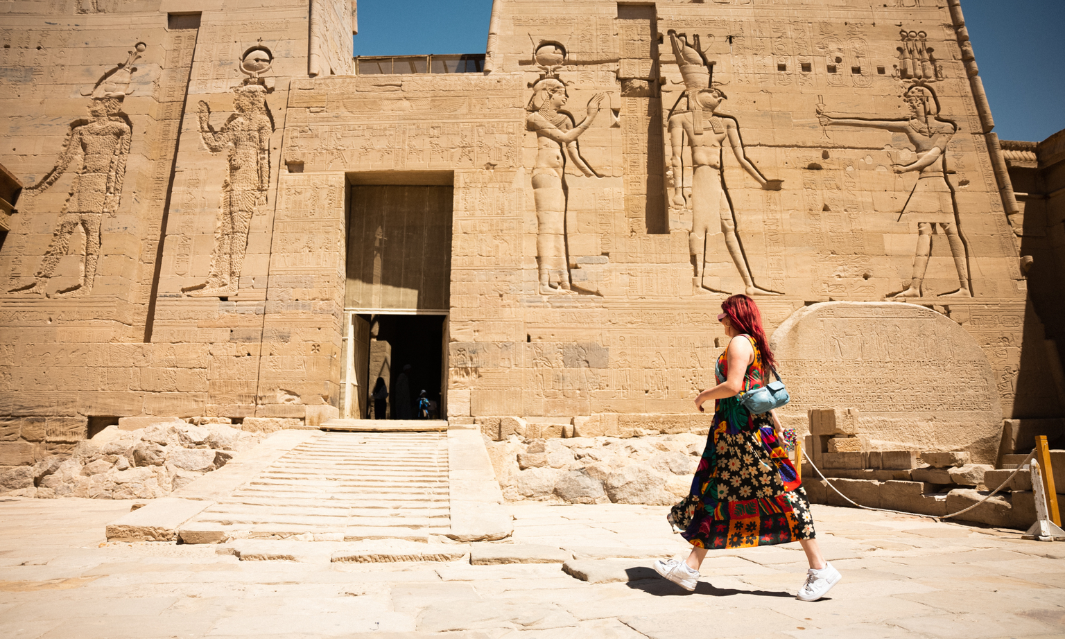 A person with red hair walks past an ancient Egyptian temple with large carvings on the walls under a clear blue sky.