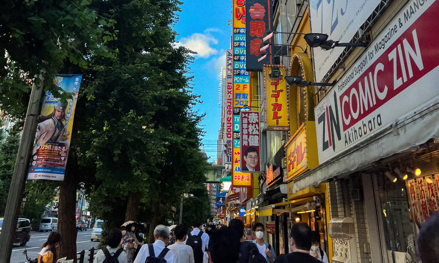 A bustling street in Akihabara, Tokyo, with colorful signs, shops, and people walking under a clear blue sky.